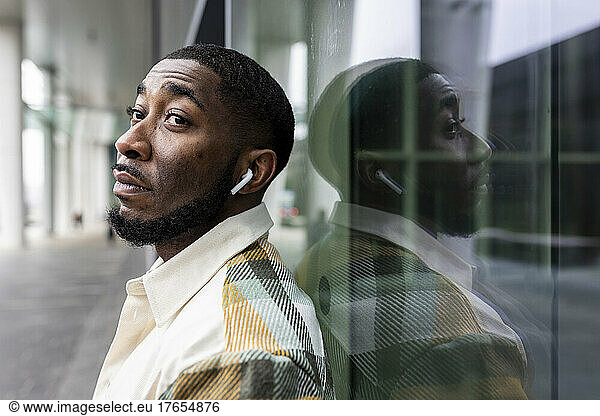 Man wearing wireless in-ear headphones leaning on glass wall