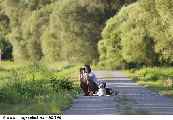 Man with a dog crouching on a road and observing something with binoculars  Wustermark  Brandenburg  Germany  Europe