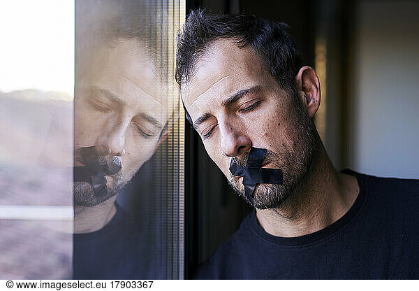 Man with adhesive tape in cross shape covering mouth leaning on window