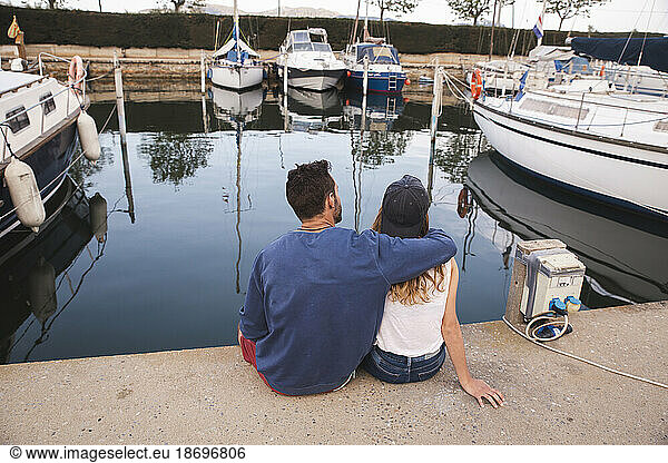 Man with arm around girlfriend sitting on jetty at harbor