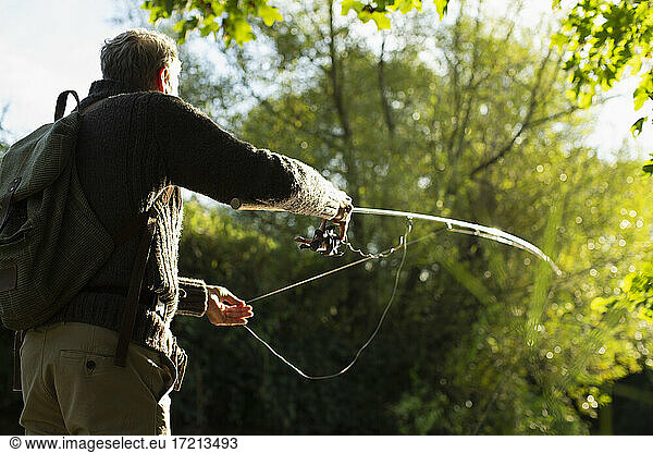 Man with backpack casting fly fishing pole below sunny trees