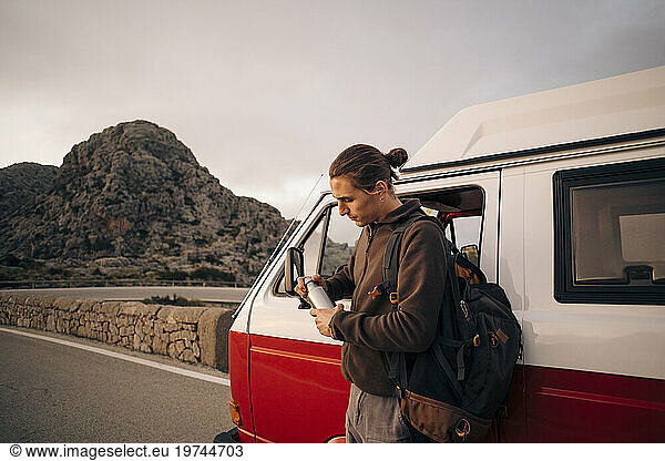Man with backpack opening water bottle while standing near van on vacation