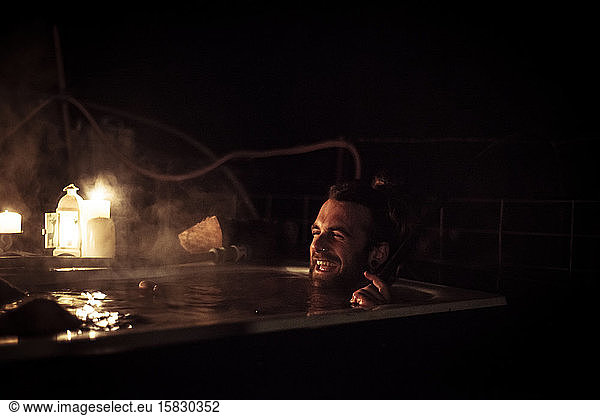 man with beard laughs in steamy outdoor bathtub with candles