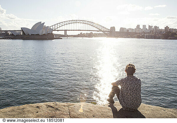 Man with bun  sitted on edge of water  staring at the Sydney Harbour