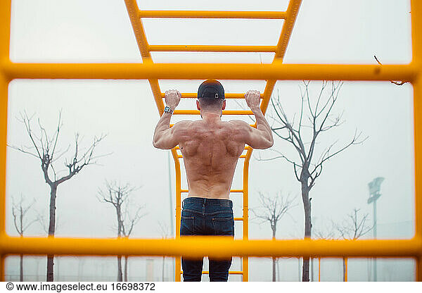 man with cap doing pull-ups in a calisthenics park. back photo
