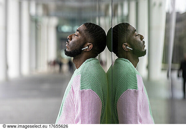 Man with eyes closed listening music through wireless in-ear headphones leaning on glass wall