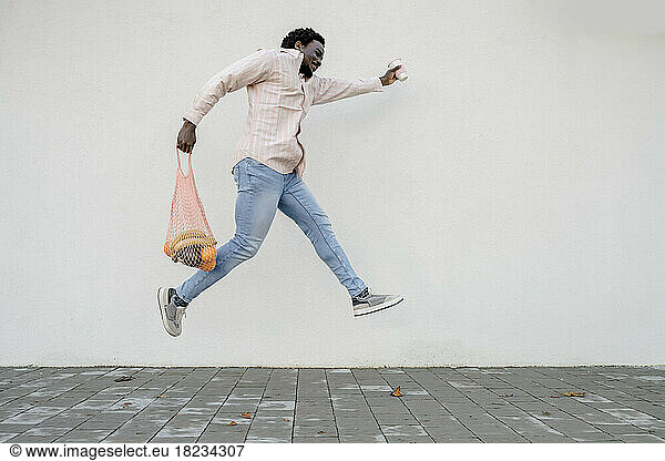 Man with grocery bag and disposable coffee cup jumping in front of white wall