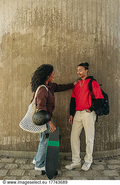 Man with hand in pocket talking with female friend holding skateboard while standing against wall