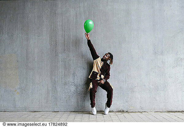 Man with hand raised holding green balloon leaning on concrete wall