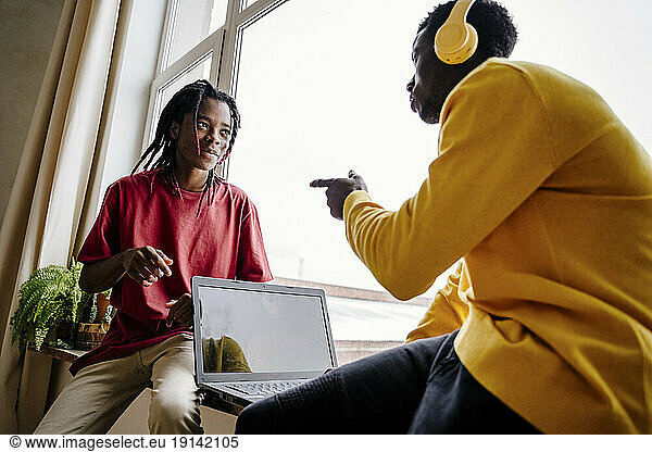 Man with laptop gesturing at friend sitting on window sill at home