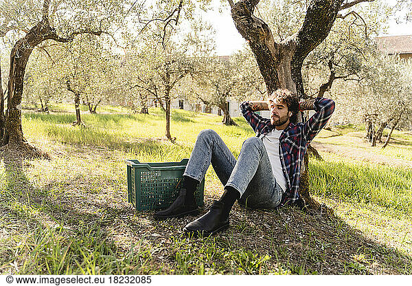 Man with plastic crate listening to music through wireless in-ear headphones leaning on tree