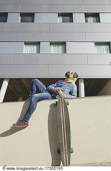 Man with pug mask lying on a wall supporting a longboard.
