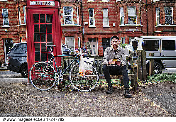 Man with smart phone sitting on bench near telephone booth at street