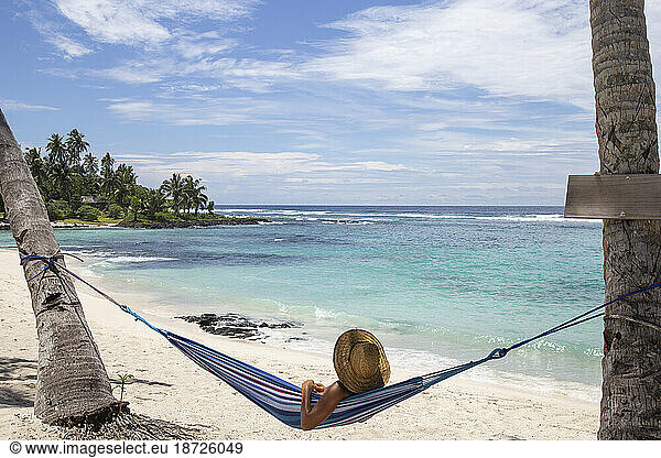 Man with straw hat  relaxing on blue hammock at sandy beach  Samoa