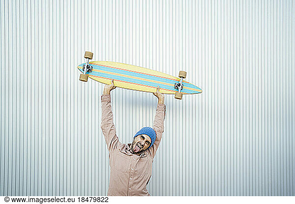 Man with tongue out holding longboard in front of white wall