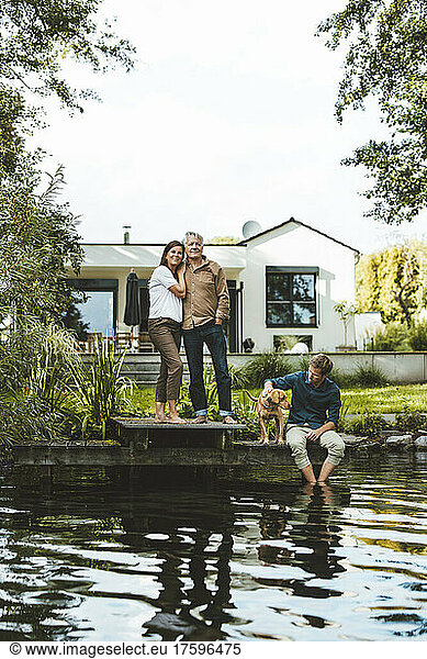 Man with woman standing by son and dog sitting by lake at backyard