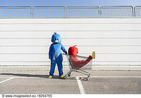 Man with woman wearing red duck costume sitting in shopping cart on sunny day
