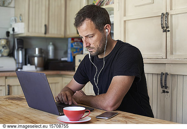 Man working from home, sitting at kitchen table, using laptop and smartphone