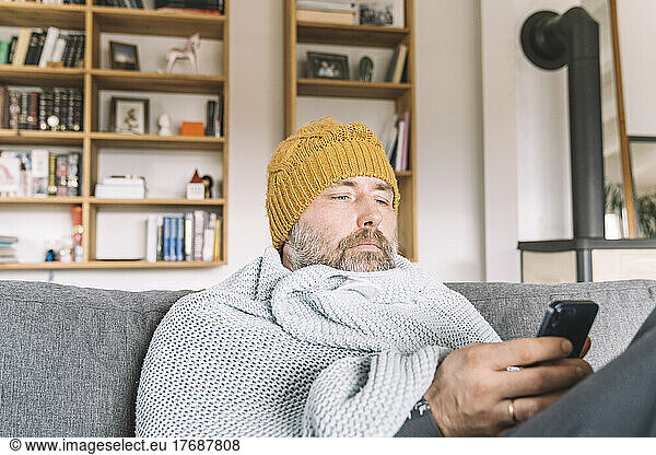 Man wrapped in blanket wearing wooly hat sitting on couch checking smartphone