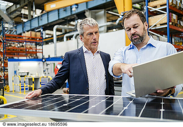 Manager holding laptop by businessman holding solar panel at factory