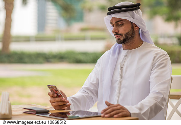 Mann aus dem Nahen Osten in traditioneller Kleidung liest Smartphone-Text im Café  Dubai  Vereinigte Arabische Emirate