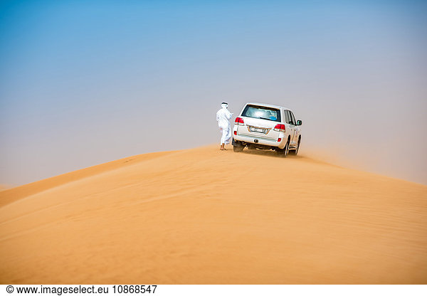 Mann aus dem Nahen Osten in traditioneller Kleidung mit Geländewagen auf Wüstendüne geparkt  Dubai  Vereinigte Arabische Emirate