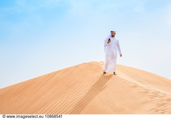 Mann aus dem Nahen Osten trägt traditionelle Kleidung mit Smartphone auf Wüstendüne  Dubai  Vereinigte Arabische Emirate