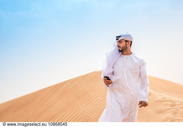 Mann aus dem Nahen Osten trägt traditionelle Kleidung mit Smartphone auf Wüstendüne  Dubai  Vereinigte Arabische Emirate