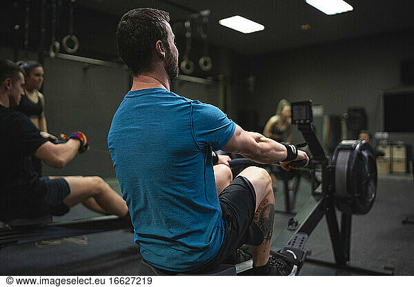 Mann benutzt Rudergerät mit Menschen  die im Hintergrund im Fitnessstudio trainieren