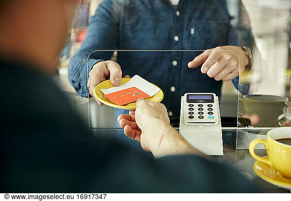 Mann übergibt Kreditkarte und Rechnung an Kellner hinter Plastikblende in Cafe
