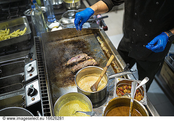Mann bestreut Fleisch mit Salz  während er am Gasherd in einer Großküche steht