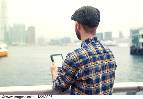 Mann hält digitales Tablett in der Hand und schaut weg  Blick auf Hafen  Rückansicht  Hongkong  China  Ostasien