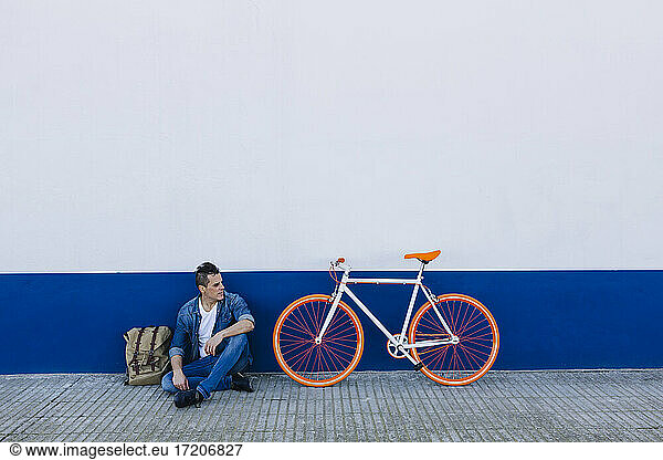 Mann in Freizeitkleidung auf dem Gehweg sitzend mit dem Fahrrad gegen die Wand