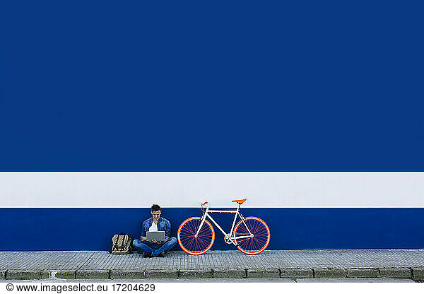 Mann in Freizeitkleidung mit Laptop auf Gehweg mit Fahrrad gegen blaue Wand
