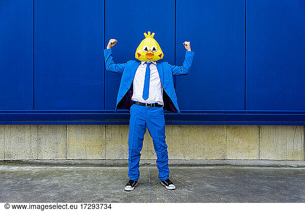 Mann in leuchtend blauem Anzug und Vogelmaske  der vor einer blauen Wand Muskeln anspannt