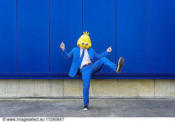 Mann in leuchtend blauem Anzug und Vogelmaske steht auf einem Bein vor einer blauen Wand