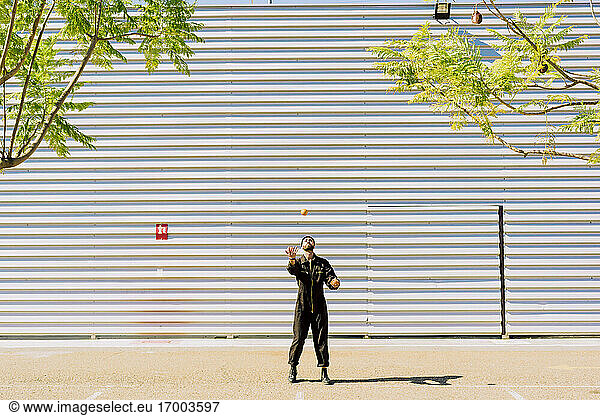Mann in schwarzem Overall jongliert mit Orangen vor einem Industriegebäude