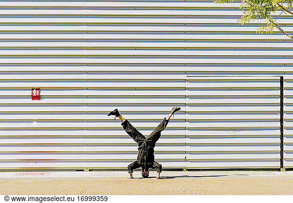 Mann in schwarzem Overall macht Handstand vor einem Industriegebäude
