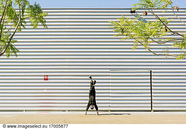 Mann in schwarzem Overall macht Handstand vor einem Industriegebäude