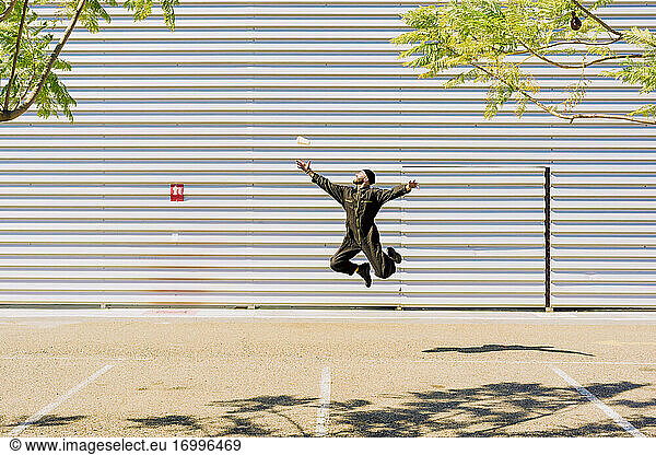 Mann in schwarzem Overall springt vor einem Industriegebäude in die Luft