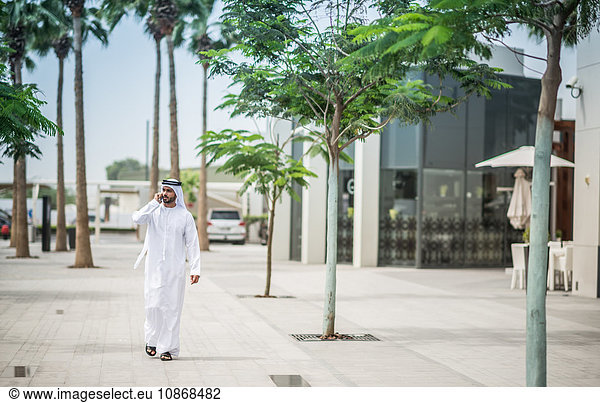 Mann in traditioneller Kleidung aus dem Nahen Osten läuft auf der Straße und spricht per Smartphone  Dubai  Vereinigte Arabische Emirate