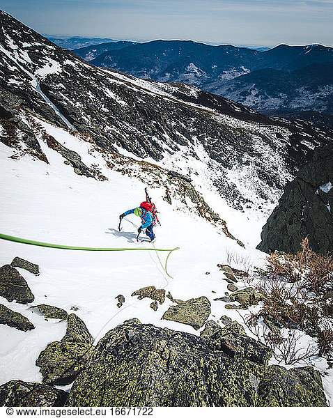 Mann klettert mit Kletterseil und Skiern eine steile Schneerinne hinauf