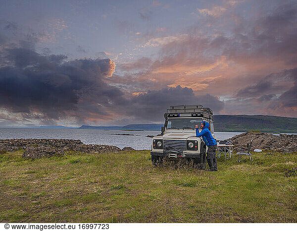 Mann lehnt sich an einen Geländewagen  der in der bewölkten Abenddämmerung an der isländischen Küste geparkt ist