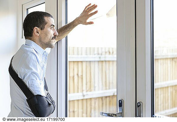Mann mit Armschlinge schaut durch ein Fenster  während er zu Hause steht