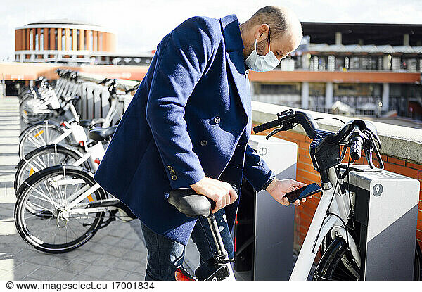 Mann mit Gesichtsmaske entriegelt an einem sonnigen Tag sein Fahrrad mit dem Smartphone