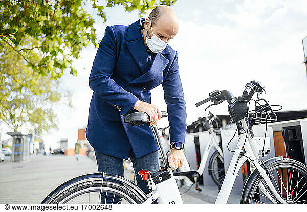 Mann mit Gesichtsschutz  der einen elektrischen Fahrradsattel einstellt  während er an einem sonnigen Tag auf einem Fußweg in der Stadt steht