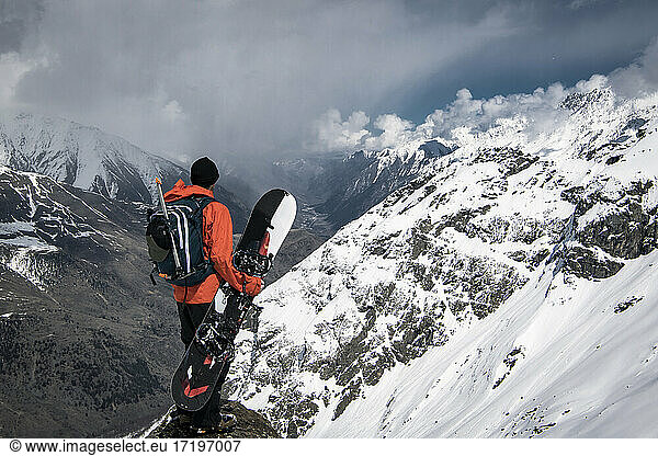 Mann mit Snowboard mit Blick auf Blick von schneebedeckten Berg gegen bewölkten Himmel