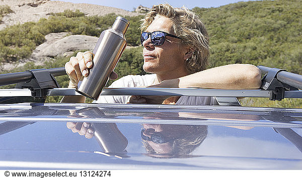Mann mit Sonnenbrille trinkt Wasser während er sich auf Auto stützt
