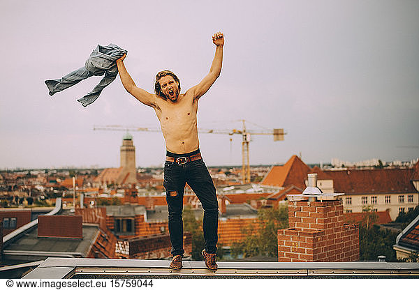 Mann ohne Hemd hält Hemd in voller Länge mit erhobenen Armen und steht auf der Stützmauer der Terrasse
