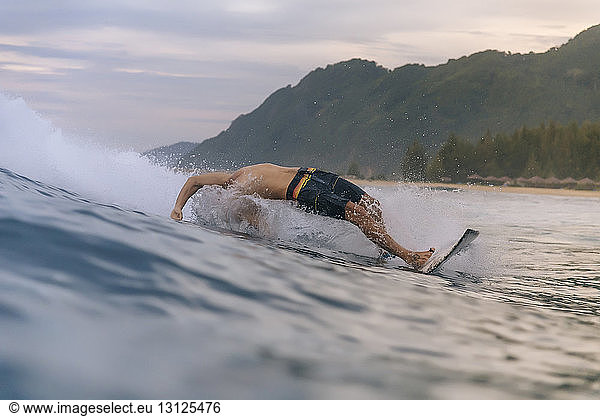 Mann ohne Hemd surft bei Sonnenuntergang auf dem Meer vor bewölktem Himmel